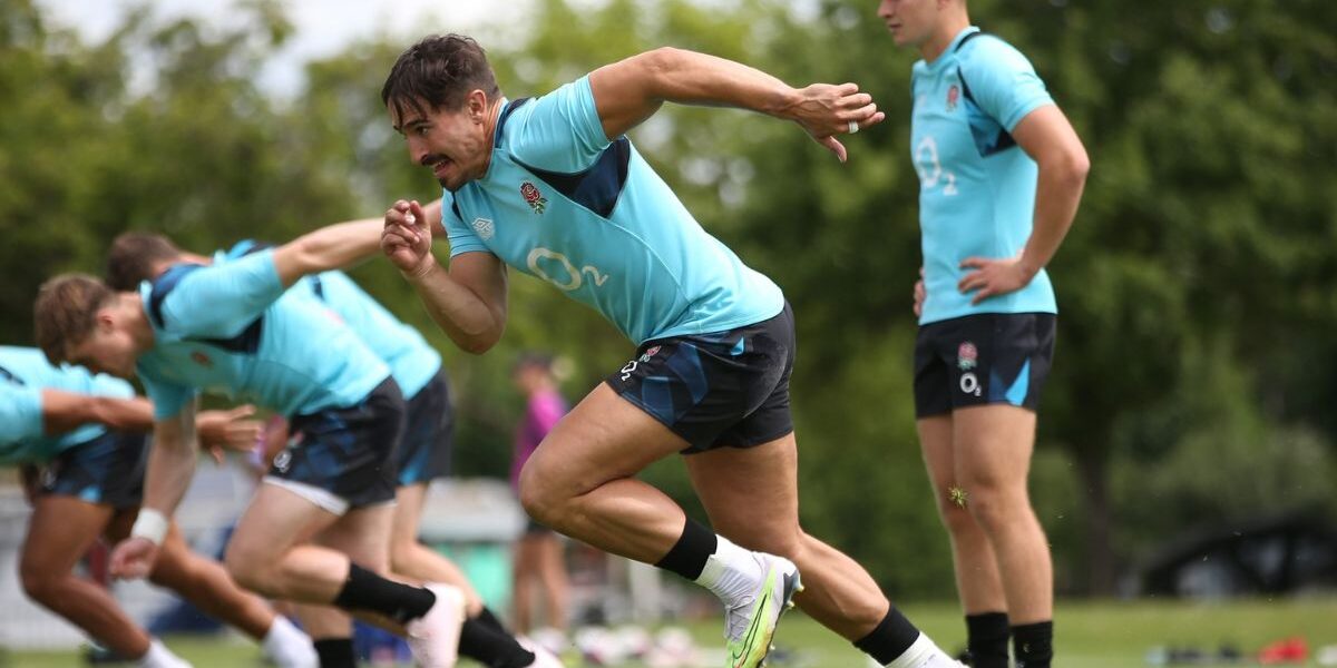 The Image Shows A Group Of Rugby Players In The Middle Of A Training Session On A Grassy Field. The Focus Is On One Player In The Foreground, Who Is Captured Mid-stride With Their Right Arm Extended Backward And Left Arm Bent Forward.